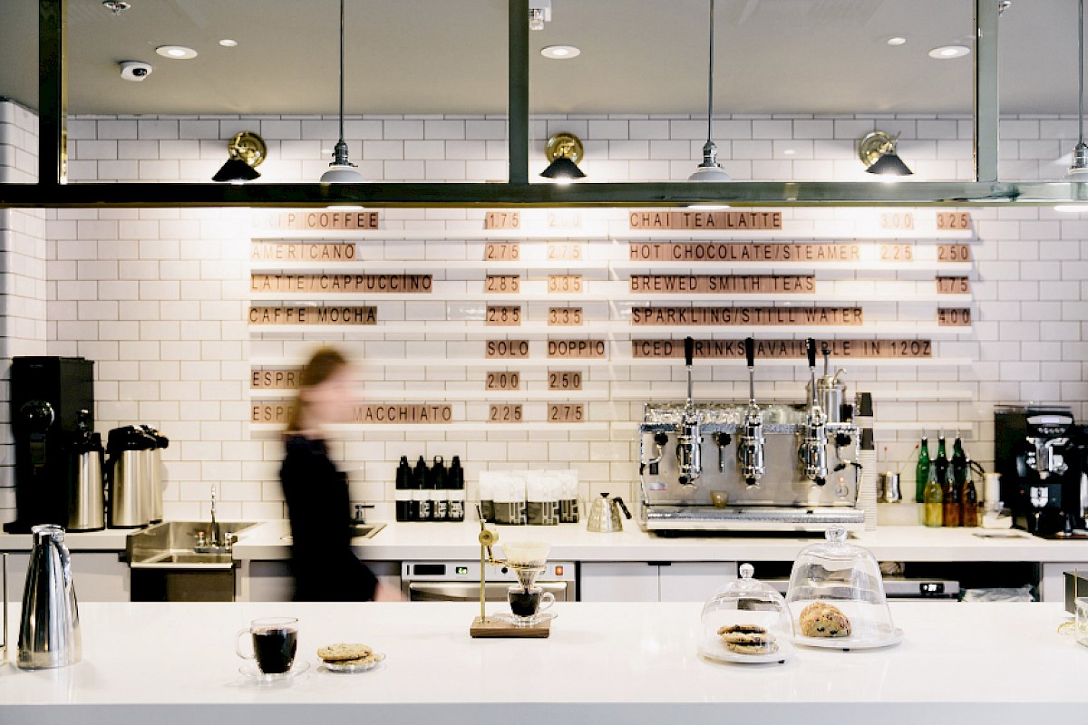 The image shows a modern coffee shop with a menu on a white tiled wall, coffee machines, pastries on the counter, and a blurred staff member.