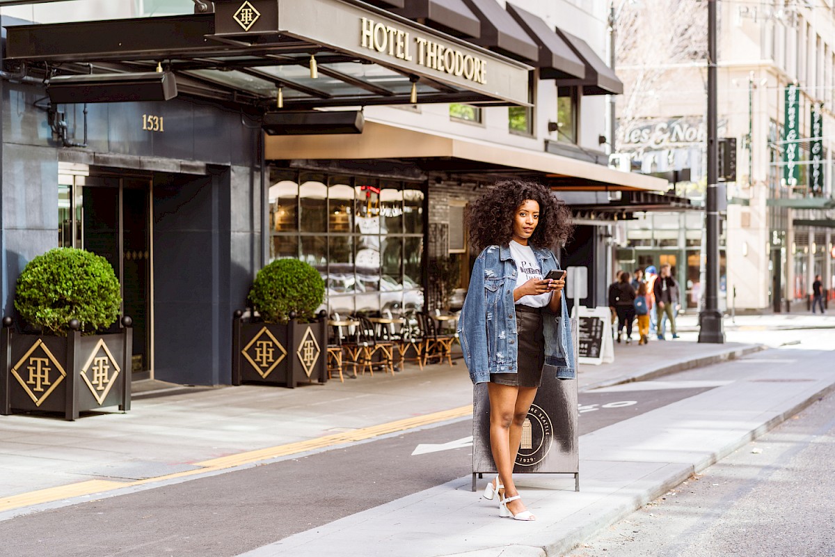 A person stands at a street corner with a phone, near Hotel Theodore. The setting is urban with modern elements and outdoor seating.
