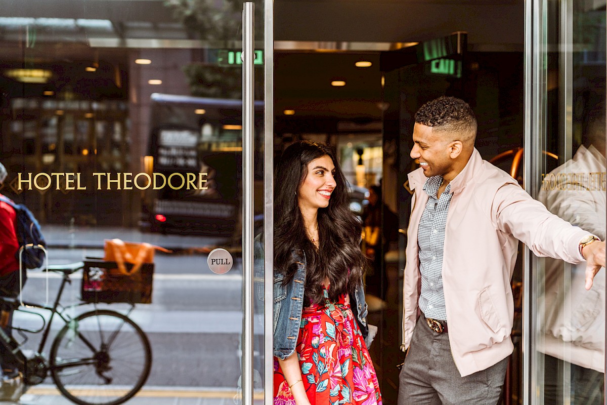 A man and woman are happily exiting Hotel Theodore, with bicycles and pedestrians visible outside on the street.