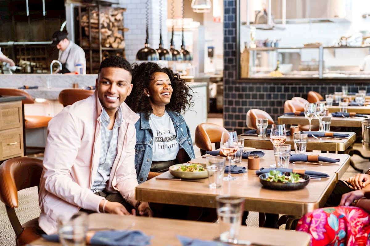 Two people are seated at a restaurant table with dishes and drinks, enjoying a meal. The background shows a bustling kitchen area.