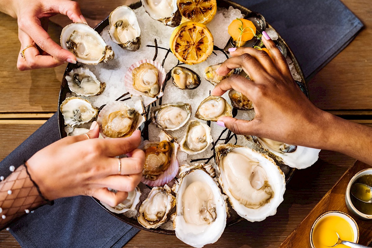 The image shows a group of people sharing a platter of oysters with lemon halves on a table with a condiments tray.