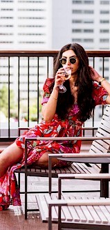 A person in a red floral dress and sunglasses sits on a balcony chair, sipping a drink with a cityscape in the background.