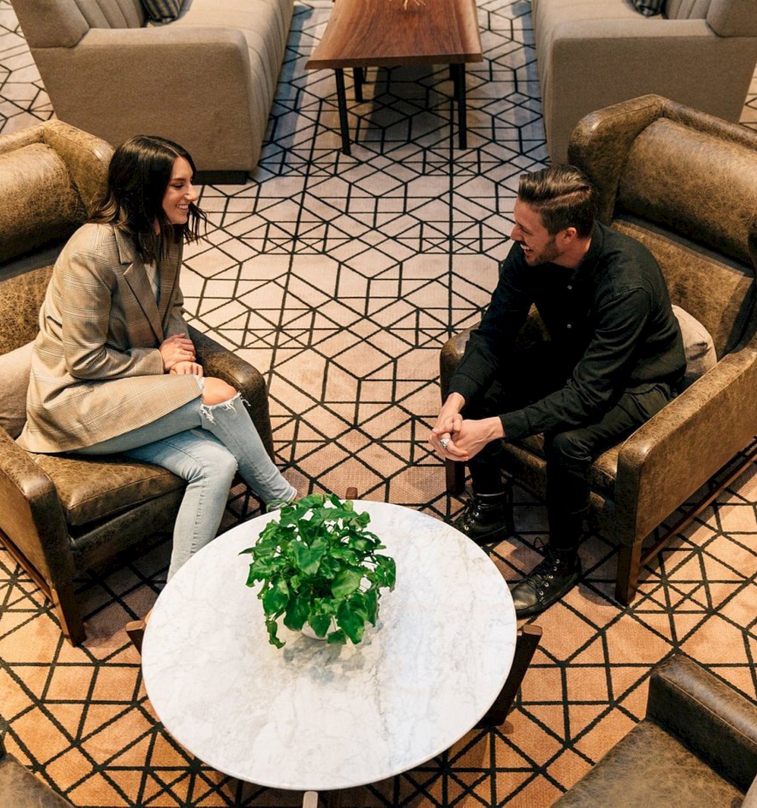 Two people sit in armchairs facing each other, with a round table and a plant between them in a stylish room.