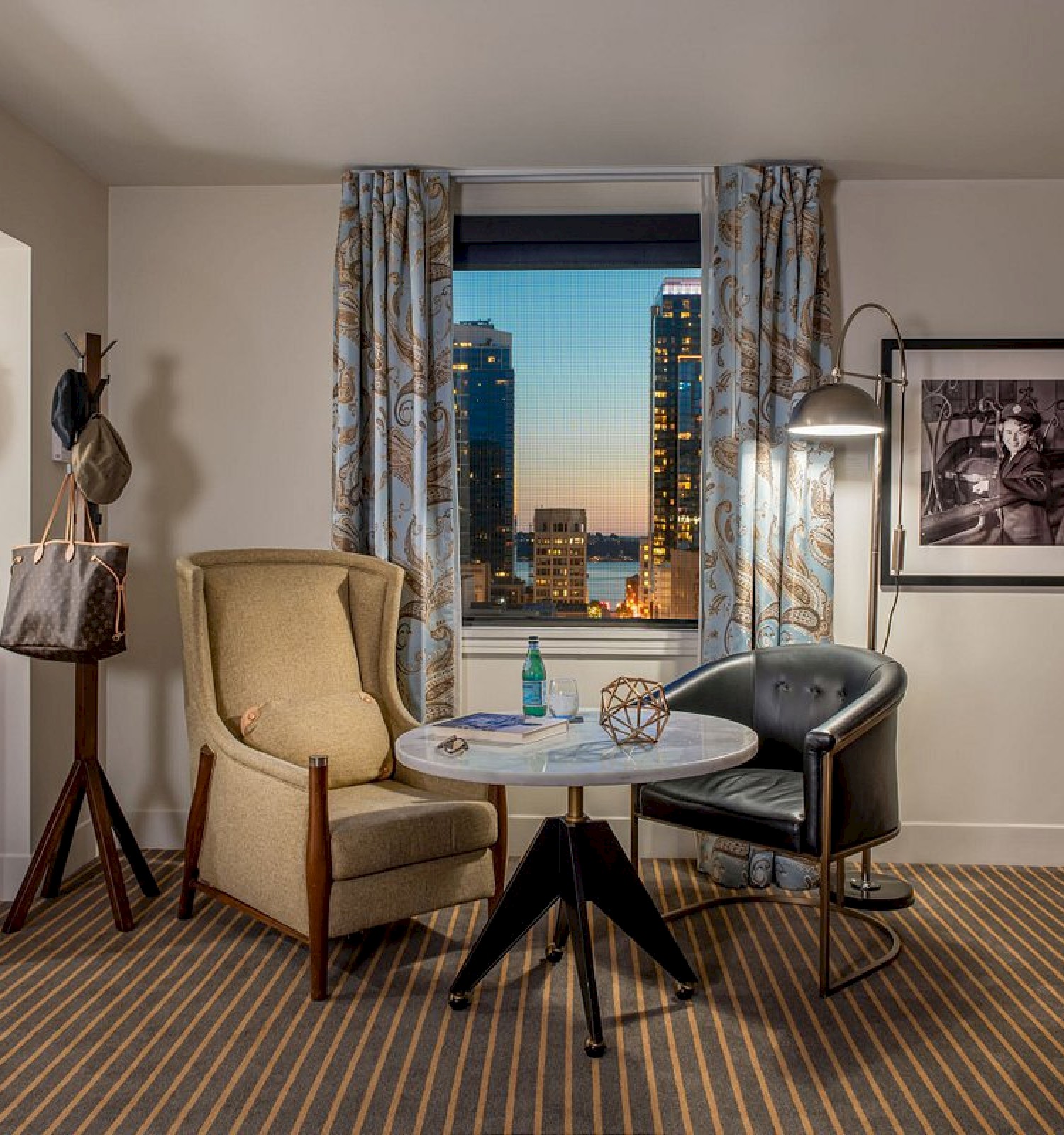 A hotel room with chairs, a small table, minibar, wall mirror, photo, lamp, and a window view of the city skyline at sunset.