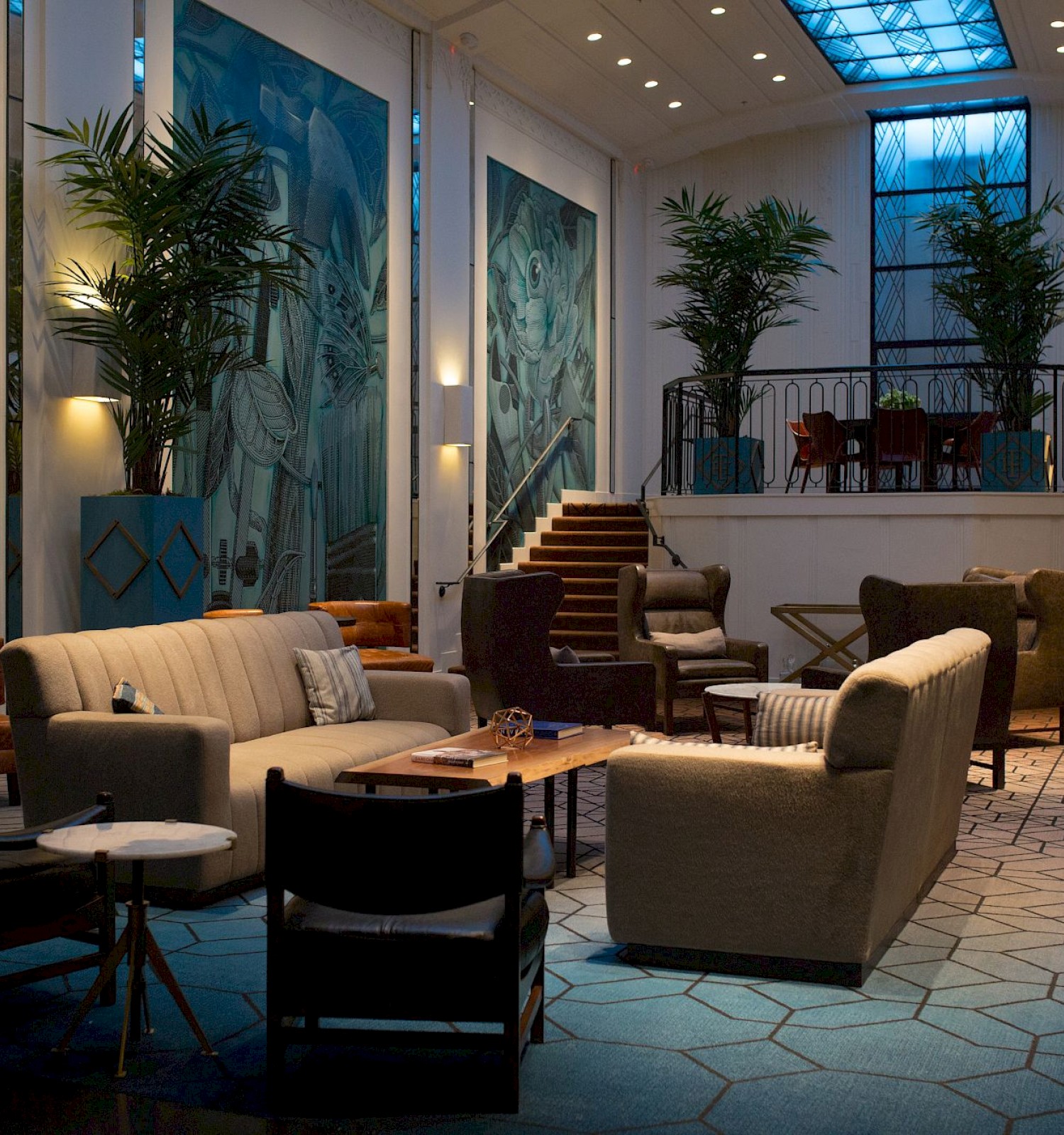 A stylish lounge with sofas, chairs, plants, and artistic walls; a staircase leads to a mezzanine with tables and chairs.