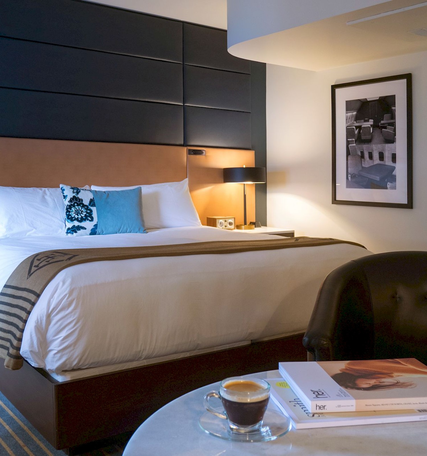 A modern hotel room with a large bed, cozy chair, and table with books and a coffee cup, creating a warm and inviting atmosphere.