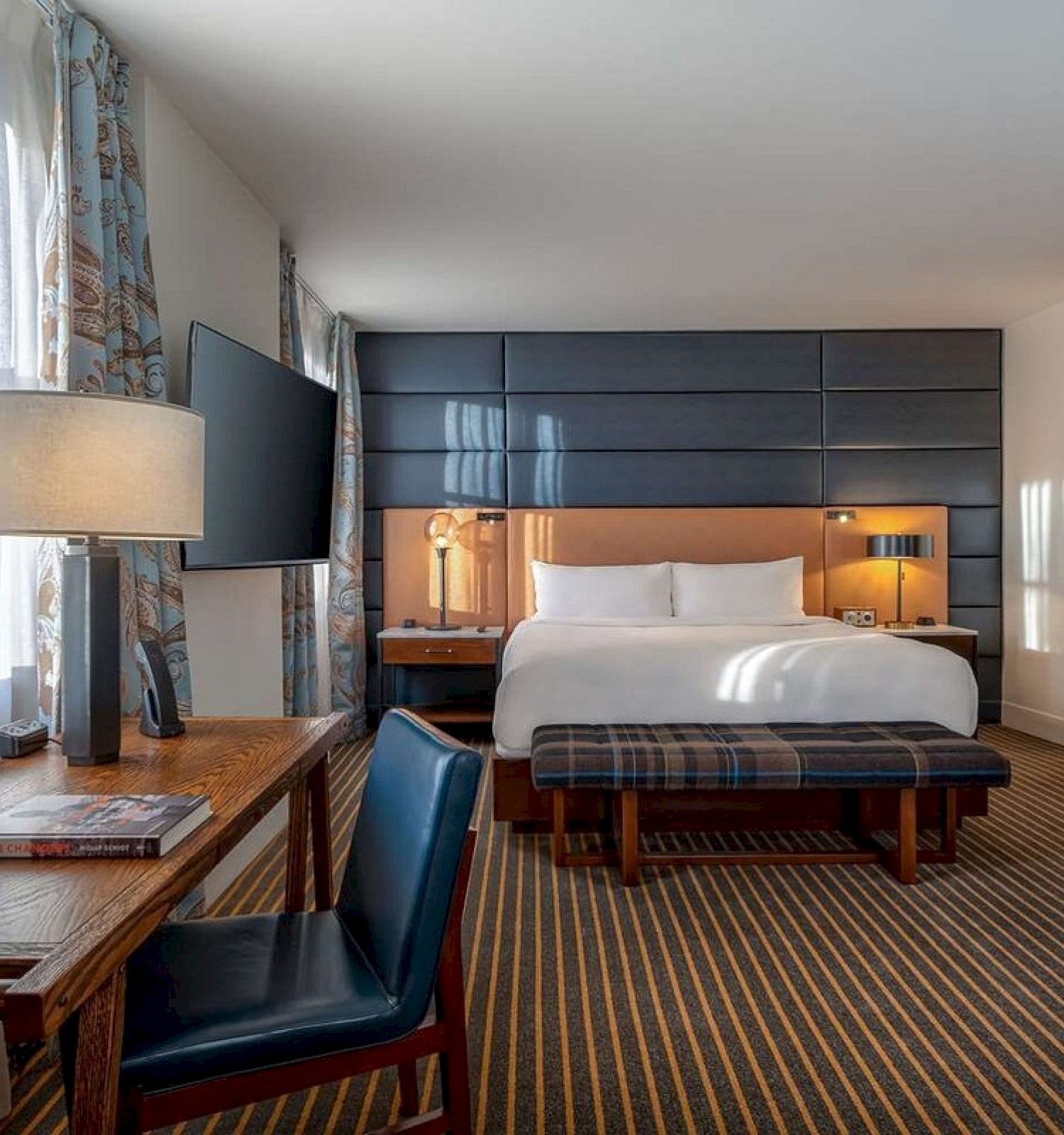 A modern hotel room with a bed, desk, chair, lamp, large TV, and artwork, featuring striped carpet and natural light from a window.