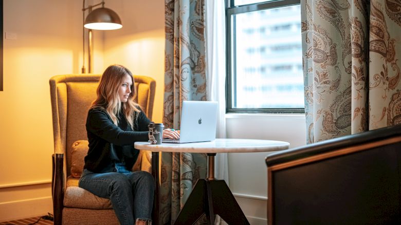 A woman is seated in a room with a laptop on a small round table, a glass of water beside her, near a window with patterned curtains.