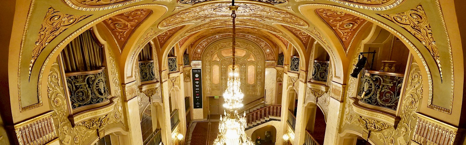 An ornate theater lobby with chandeliers, decorated ceilings, and elegant archways, featuring rich crimson carpeting and balconies.