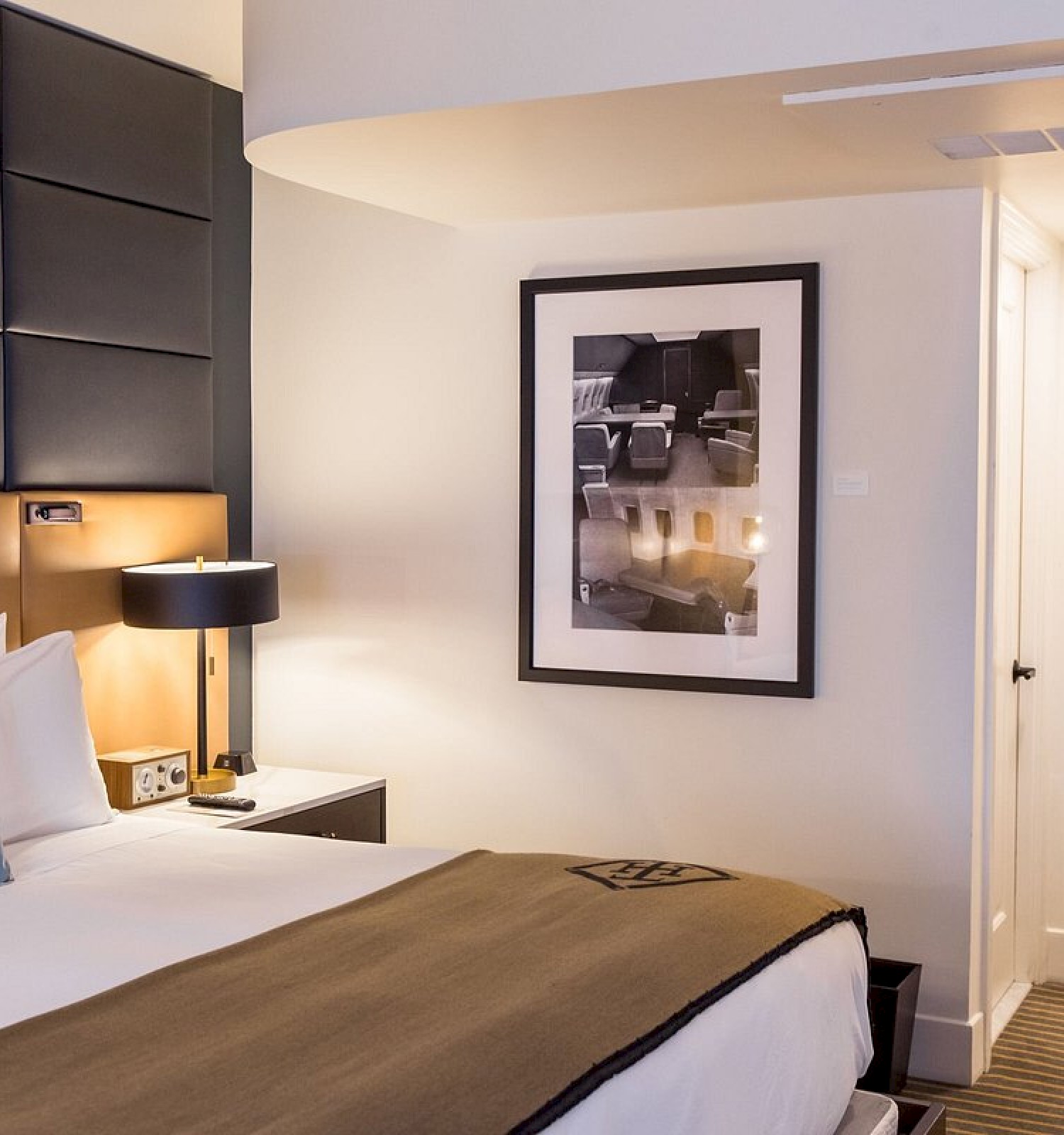 A cozy hotel room featuring a bed, pillows, wall art, a bedside table with a lamp, and a minibar with a mirror above it.