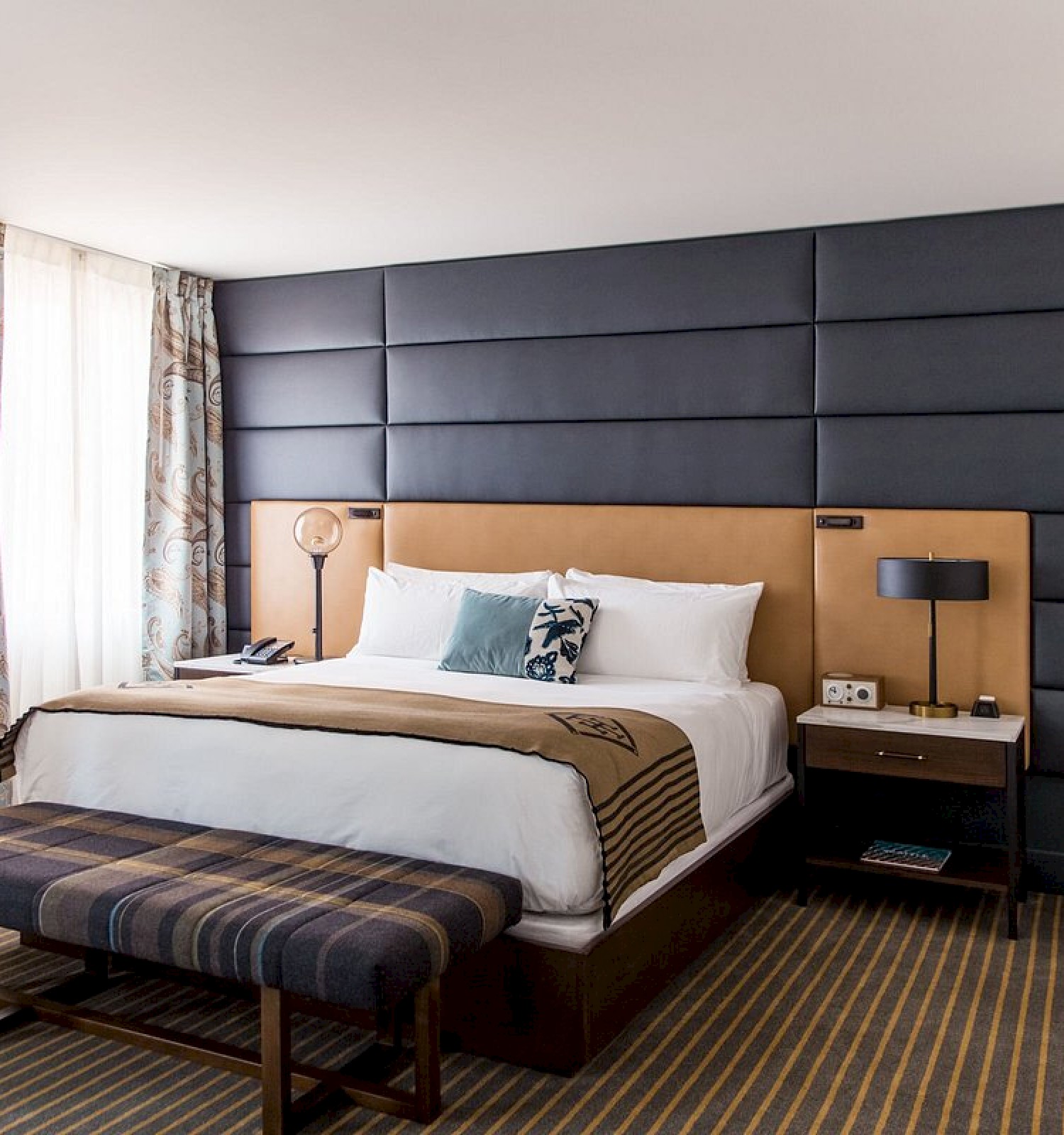 A modern hotel room with a large bed, striped bench, window with curtains, side tables, and wall-mounted TV.