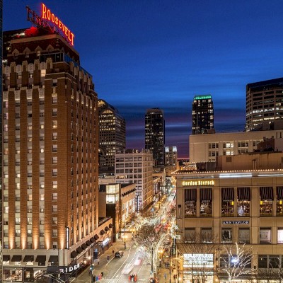 A cityscape at dusk showing illuminated buildings, including a prominent hotel, and busy streets lined with shops and offices.