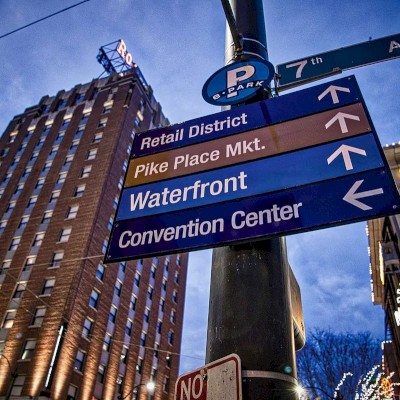 A street sign points to Retail District, Pike Place Market, Waterfront, and Convention Center against a city backdrop in the evening.