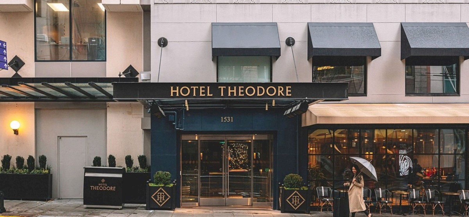 The image shows the entrance of Hotel Theodore with a person holding an umbrella, and outdoor seating to the right.