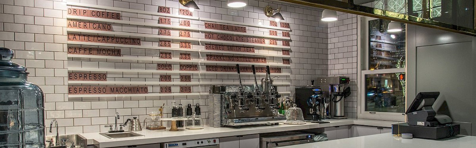 The image shows a modern coffee shop interior with a white counter, stools, an espresso machine, and a menu on a tiled wall.