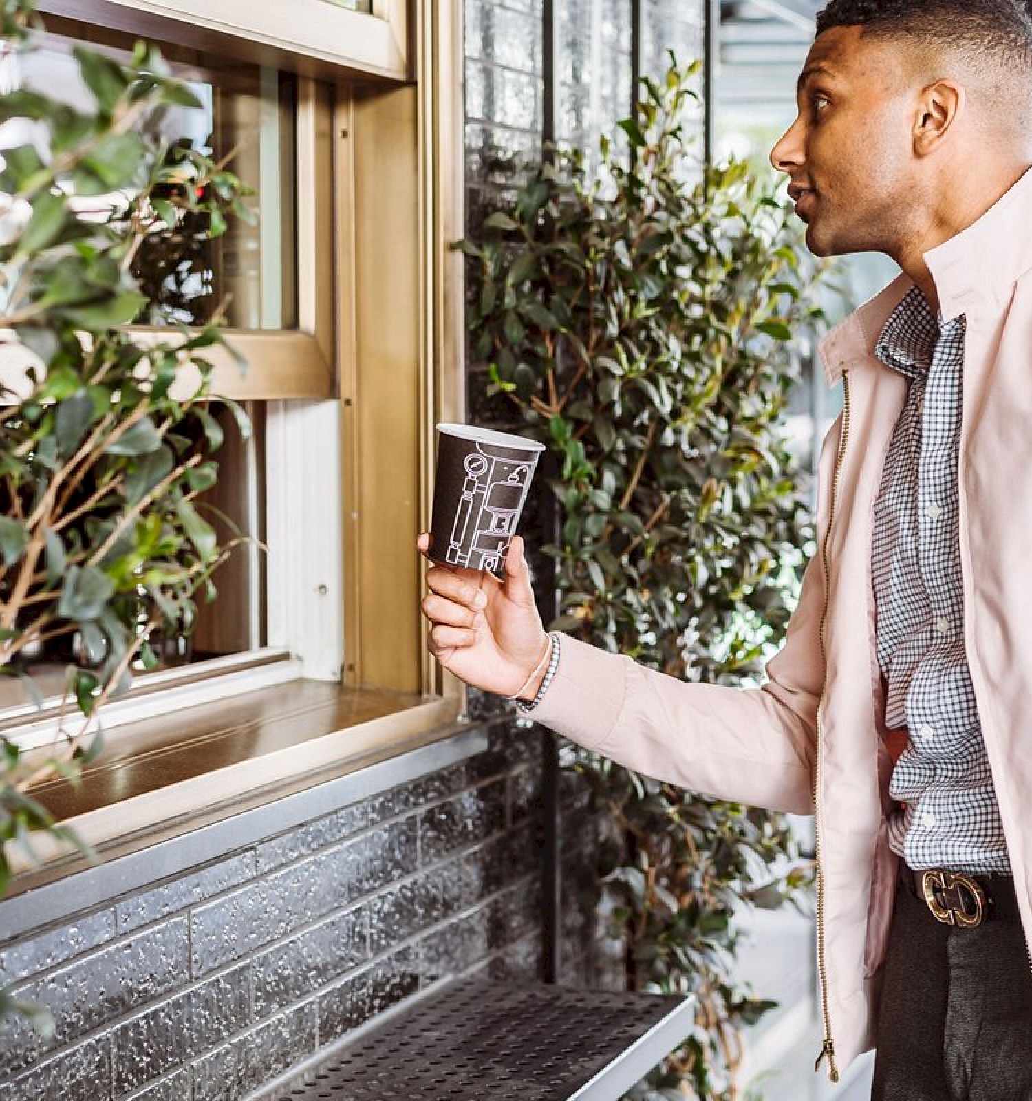 A person is standing at a window holding a cup, possibly waiting for coffee. They're wearing a light jacket and surrounded by greenery.