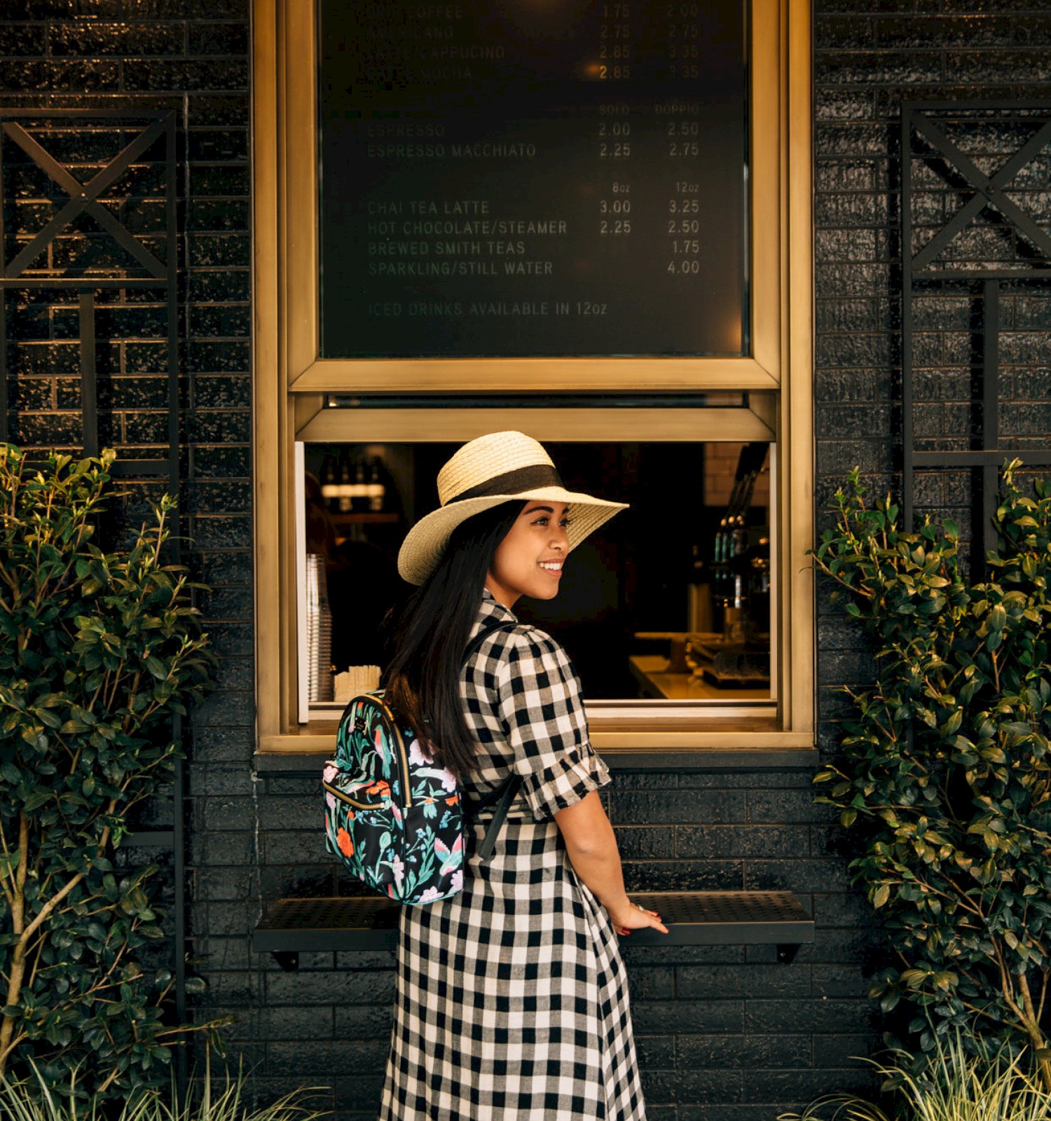 A person in a checkered dress and hat stands in front of a service window, surrounded by plants, with a menu board above.