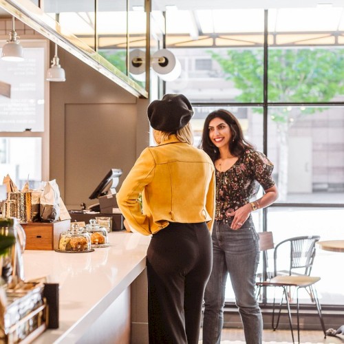 Two people are conversing at a café counter, with small tables visible near large windows in the background.
