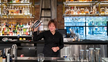 A bartender in a black outfit is shaking a cocktail shaker behind a bar with bottles and glasses on shelves.