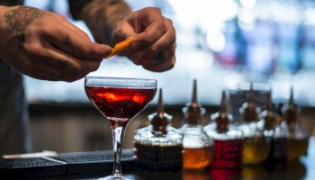 A person is preparing a cocktail at a bar, garnishing it with an orange peel surrounded by bottles of mixers.