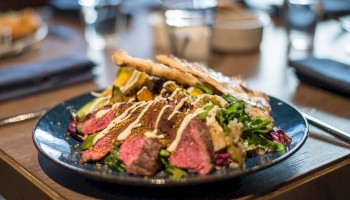 The image shows a plate of sliced steak with greens, drizzled sauce, and crispy bread on a dining table setting.