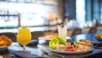 A table set with a variety of dishes, including a salad, meats, a yellow drink, and a clear drink, in a modern dining setting.