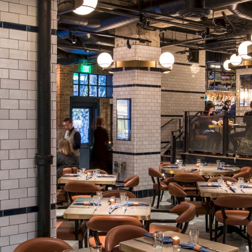 A stylish restaurant with wood tables, leather chairs, and tiled columns. It features industrial lighting and a cozy, modern atmosphere.