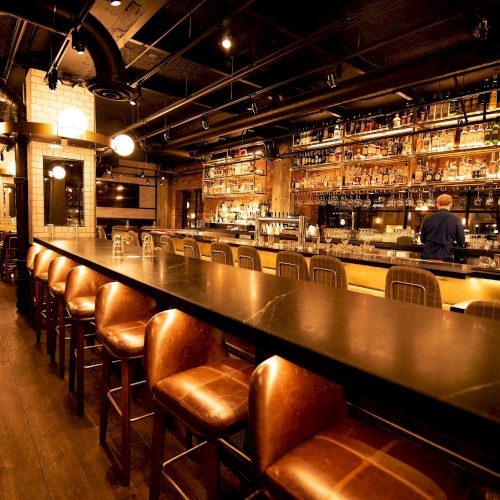 The image shows a warmly lit bar with leather stools, a long counter, and shelves filled with bottles behind. A bartender is at work.