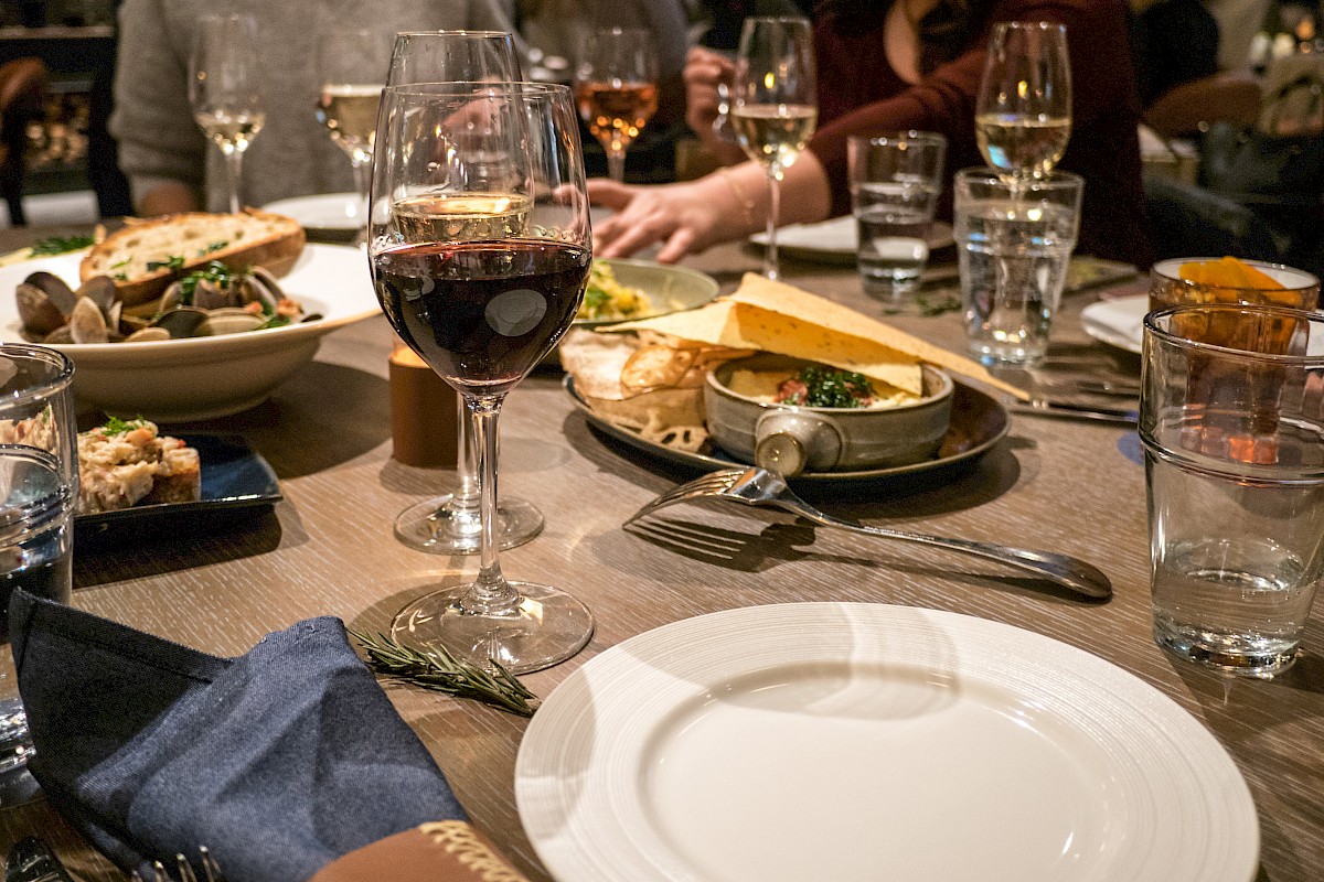 A dining table is set with plates, glasses of wine, water, and various dishes of food, with people sitting around enjoying the meal.