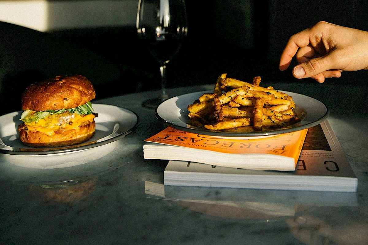 A burger and fries on plates, positioned on books, with a hand reaching for fries and a wine glass in the background, lit by sunlight.