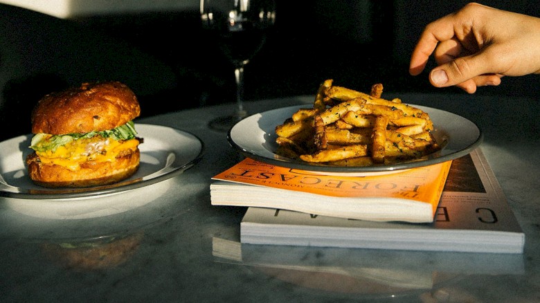 A burger and fries on plates, positioned on books, with a hand reaching for fries and a wine glass in the background, lit by sunlight.