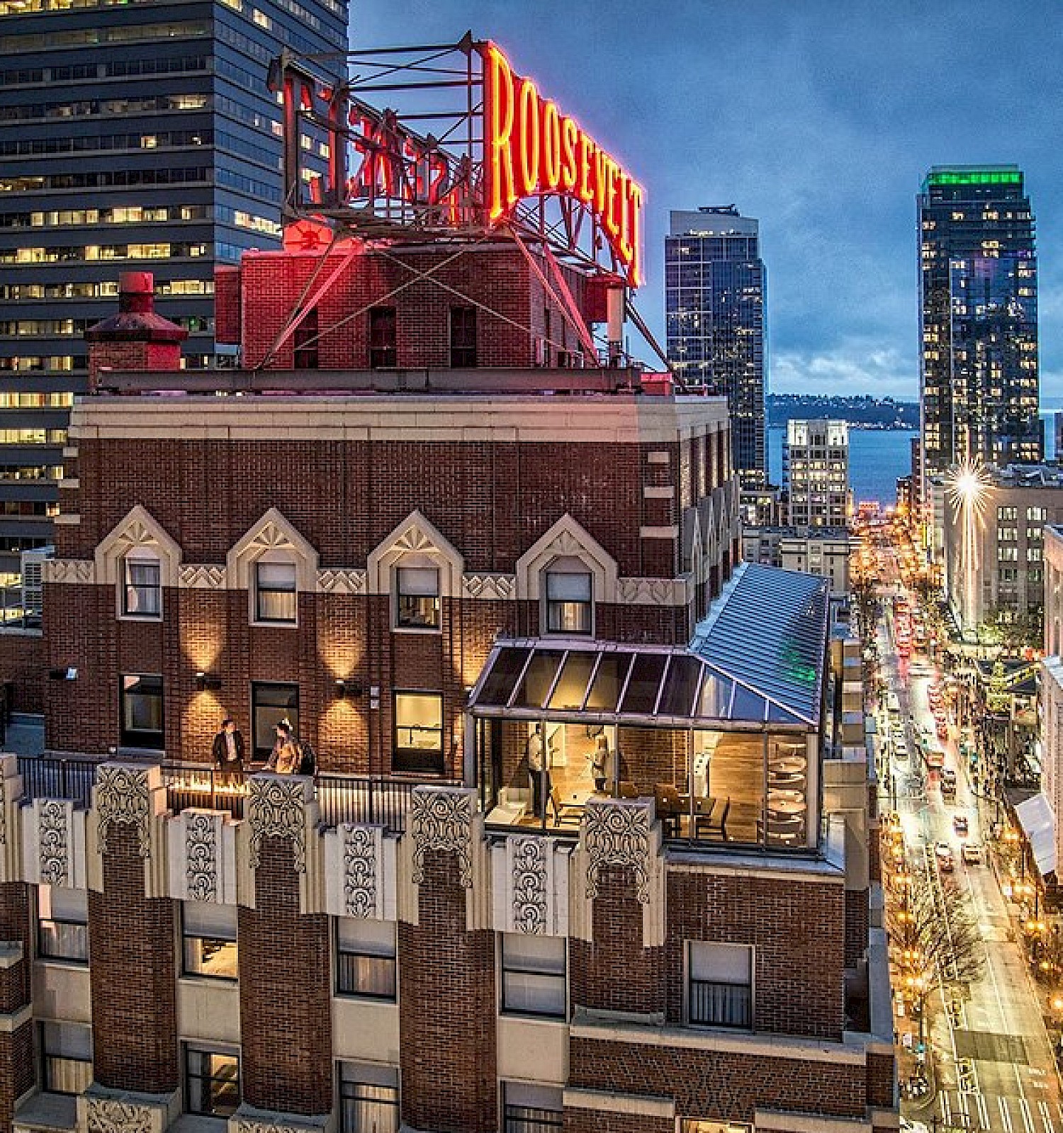 A rooftop with a neon sign in a cityscape at dusk, surrounded by tall buildings and a busy street with lights.