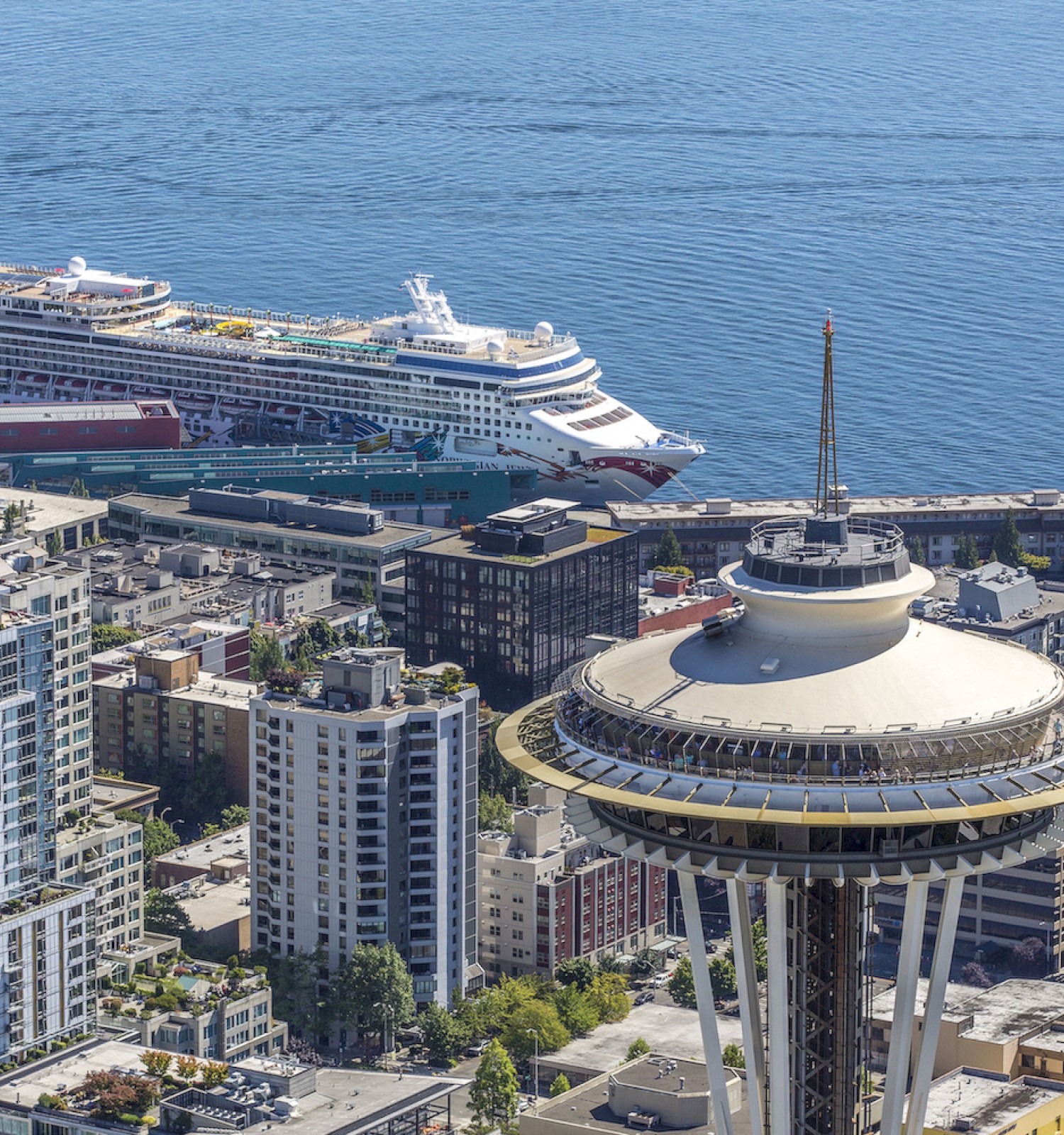 A cityscape with high-rises near a port; a large cruise ship docked beside a circular observation tower (like Seattle’s Space Needle style) above water.