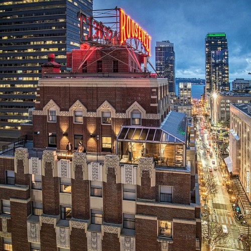 A city rooftop with brick Art Deco building, illuminated windows, and a bright street below lined with tall modern skyscrapers at dusk.