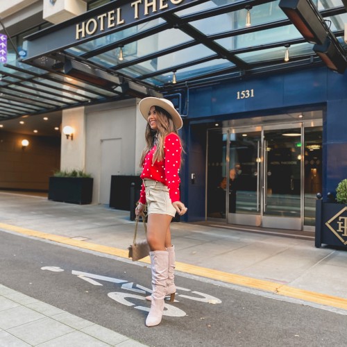 A stylish woman in a red polka-dot top and white shorts, wide-brim hat, knee-high boots, standing outside Hotel Theodore in a chic urban street scene.