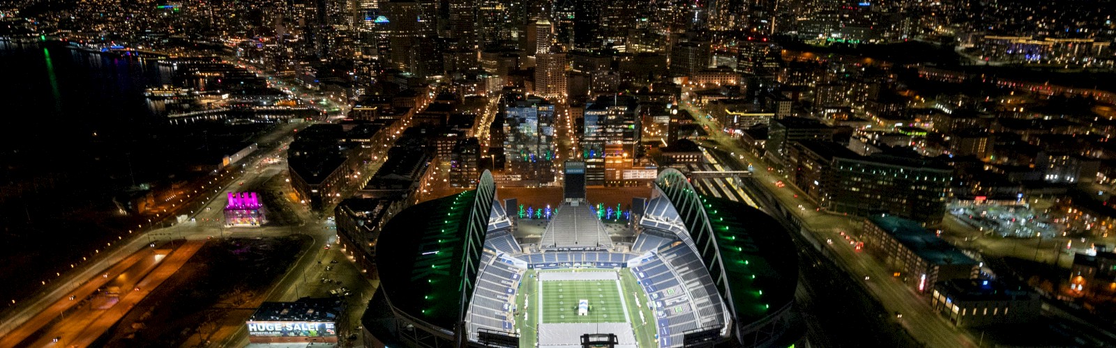 Aerial view of a glowing city at night with a lit football stadium in the center, surrounded by streets and tall buildings, shining bright.