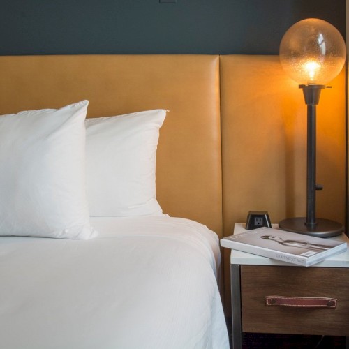 A tidy hotel bedroom scene with a bed, white pillows, a floral accent pillow, a nightstand, a glowing lamp, and a window next to the bed.
