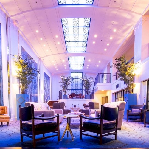 A spacious, modern hotel lobby with colorful blue and purple lighting, plants, and seating clusters arranged around small tables, creating a welcoming lounge.