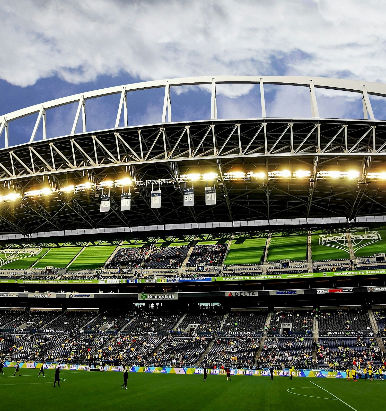 A large football stadium with a curved white roof, green seats, and players on the field under bright stadium lights.