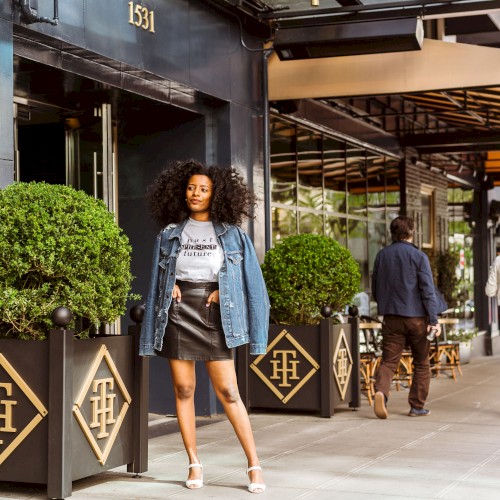 A stylish woman stands on a city sidewalk in front of a chic storefront, wearing a denim jacket, black mini skirt, and heels, with greenery and passersby around.
