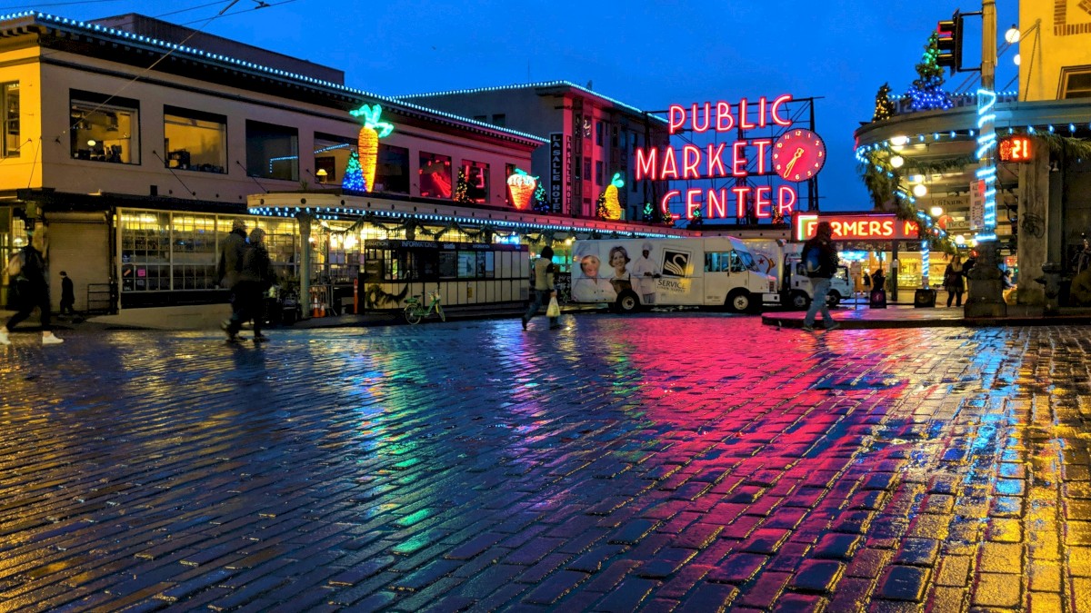 A colorful night scene at a busy market plaza with glowing signs, neon lights, and a wet cobblestone square reflecting vibrant colors, people strolling.