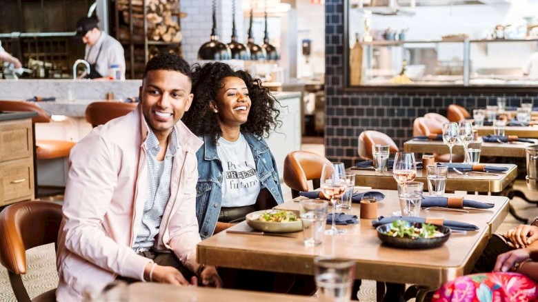 Two people smile while sitting in a restaurant with food and drinks on the table, surrounded by a cozy atmosphere and modern decor.