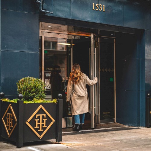 A person in a beige coat opens a glass door to a blue storefront, stepping into a chic urban entrance with potted plants flanking the door.