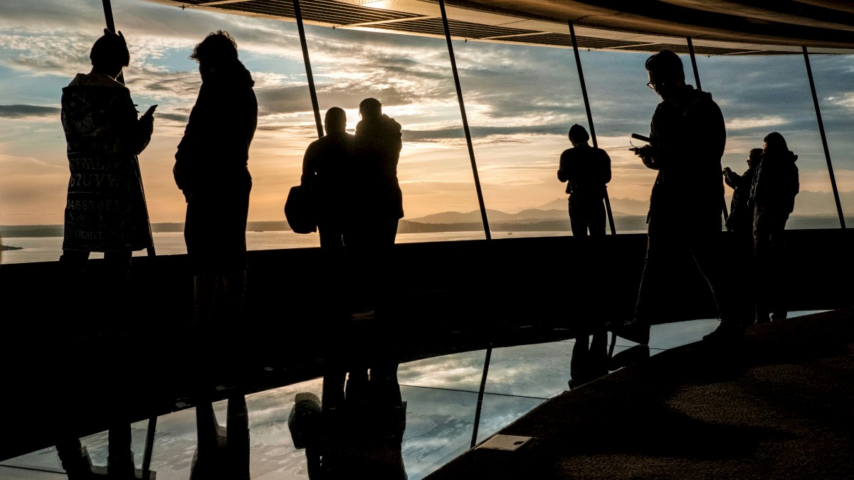 People stand and talk along a glass railing at sunset, their silhouettes reflected on the shiny floor, creating a calm, moody scene.