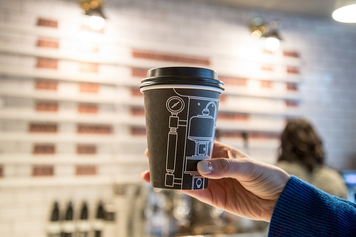 A hand holds a coffee cup with a design, in a cafe setting featuring a blurred brick and tile wall in the background.