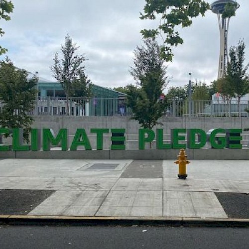 A large green sign reads “CLIMATE PLEDGE ARENA” at Seattle Center, with trees, a fire hydrant, and a cloudy sky in the background.