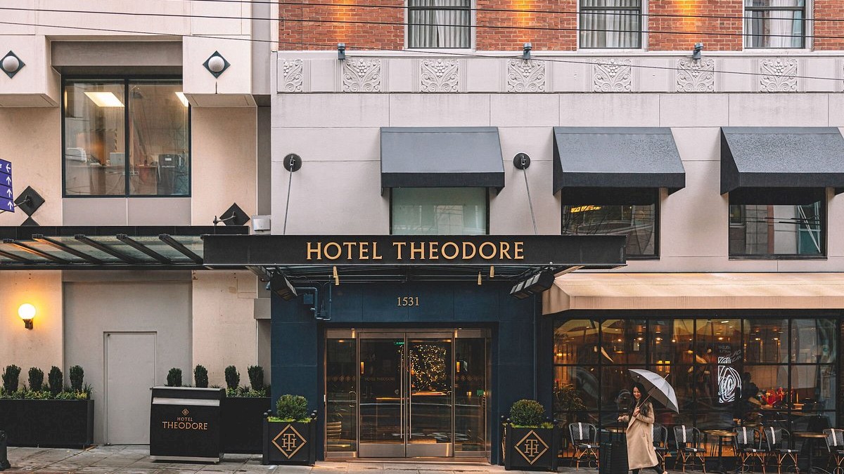 A city hotel entrance with a blue awning reading “Hotel Theodore,” glass doors, outdoor seating, and warm lights on a brick building.