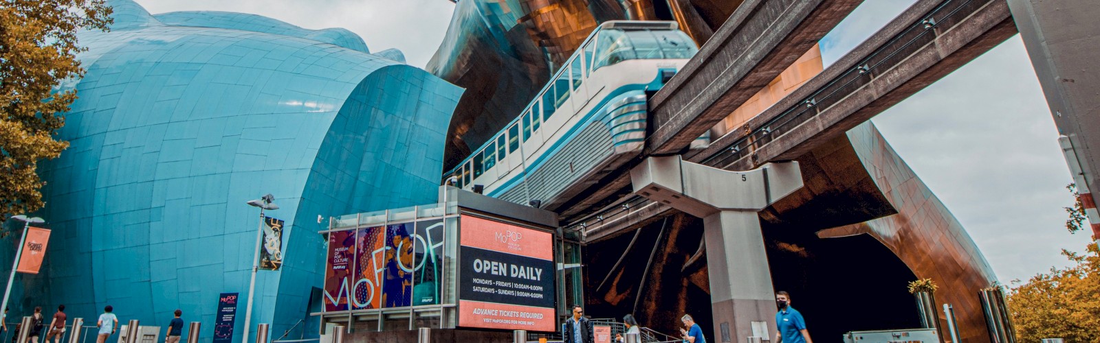 A futuristic architectural site with curving metal structures, a blue bubble-like building, an elevated monorail/tram, and people walking near signage.