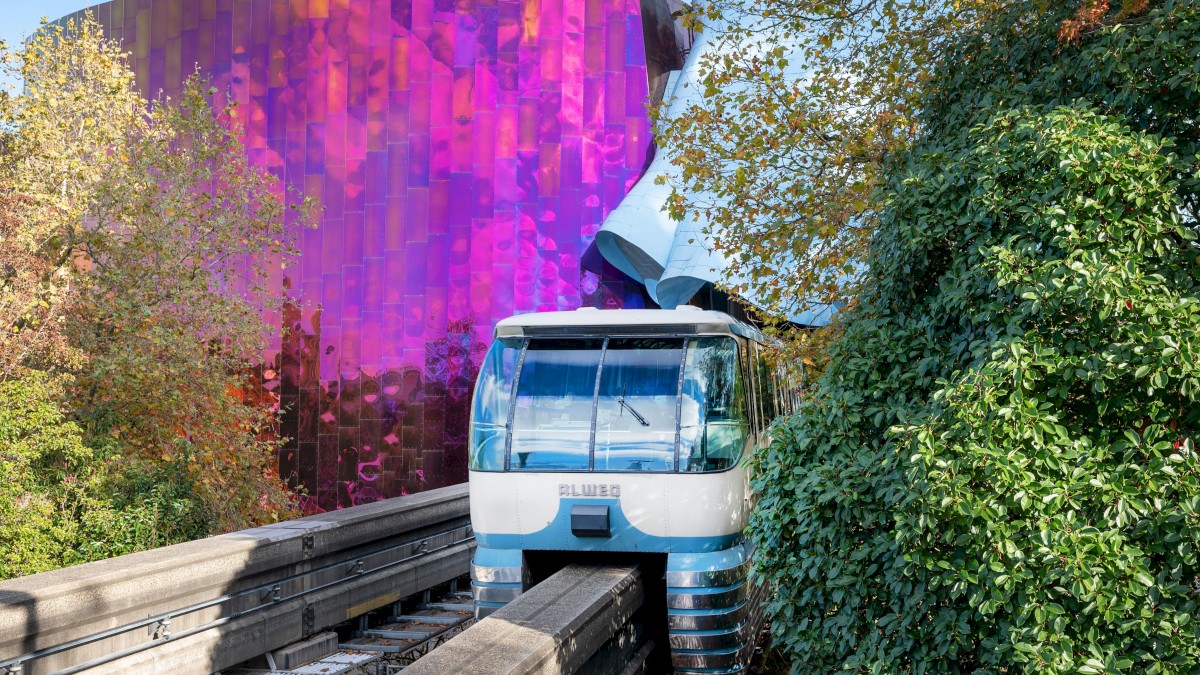 A blue and white funicular train ascends a track flanked by green foliage, with a striking purple-curtain glass building in the background.