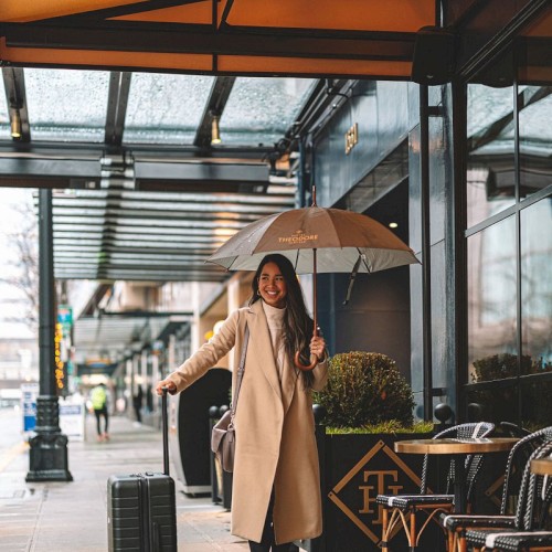 A woman in a beige coat with a suitcase and umbrella walks down a cloudy city sidewalk outside a cafe, under an orange awning.